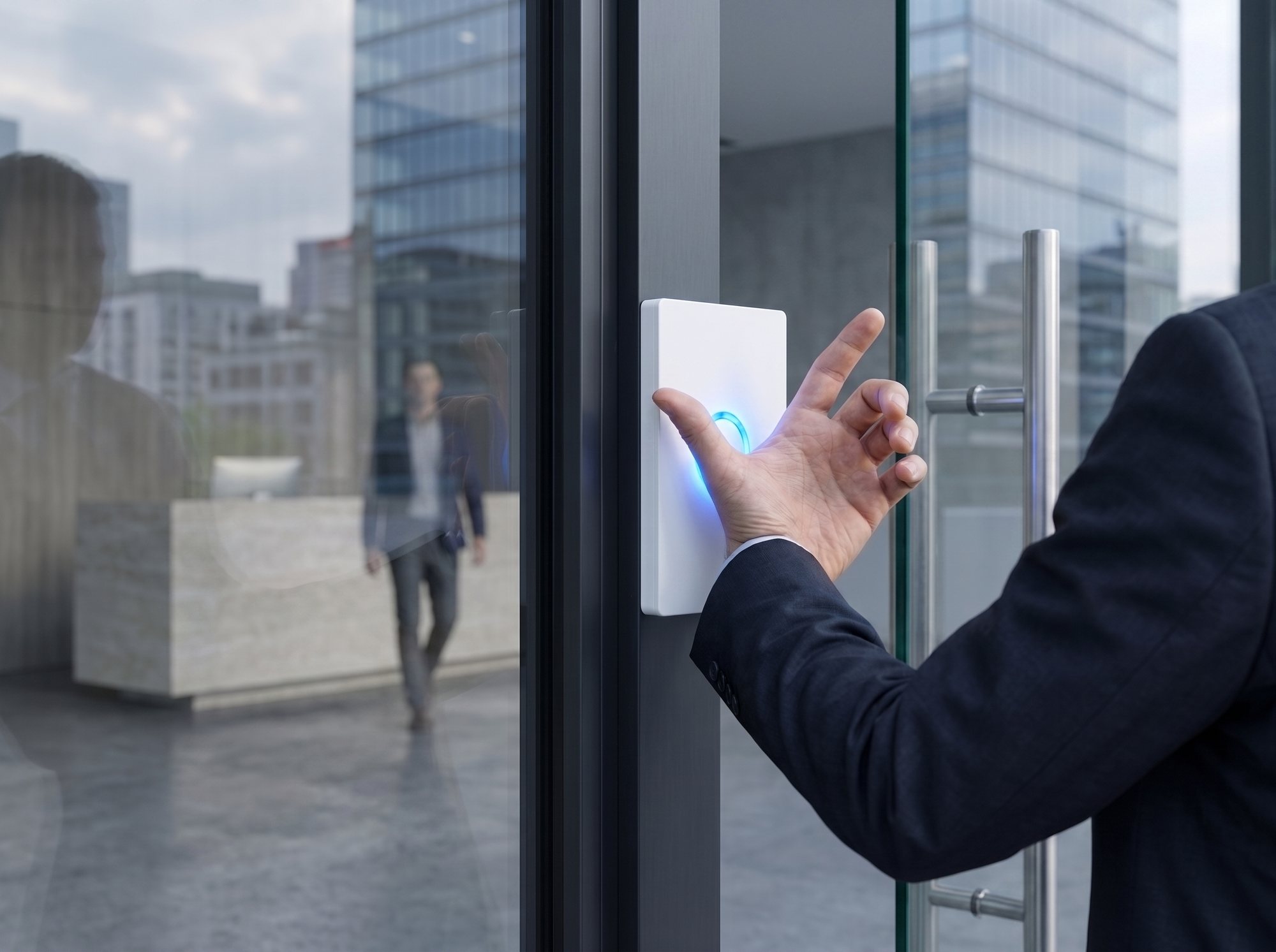 A hand held against a wall reader beside a glass office door, the reader ring lit blue